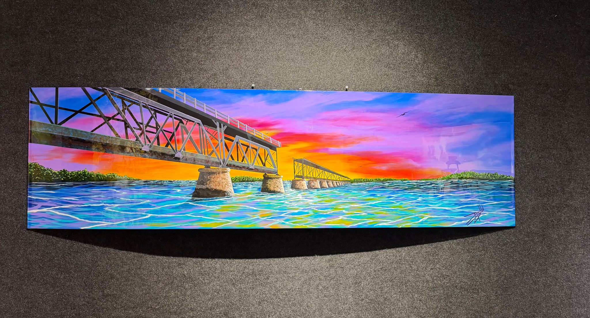 Panoramic painting of a bridge above vivid turquoise water beneath a pink and orange sunset